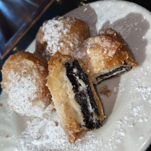 Four deep-fried Oreos dusted with powdered sugar on a white plate, one cut open revealing the cookie.
