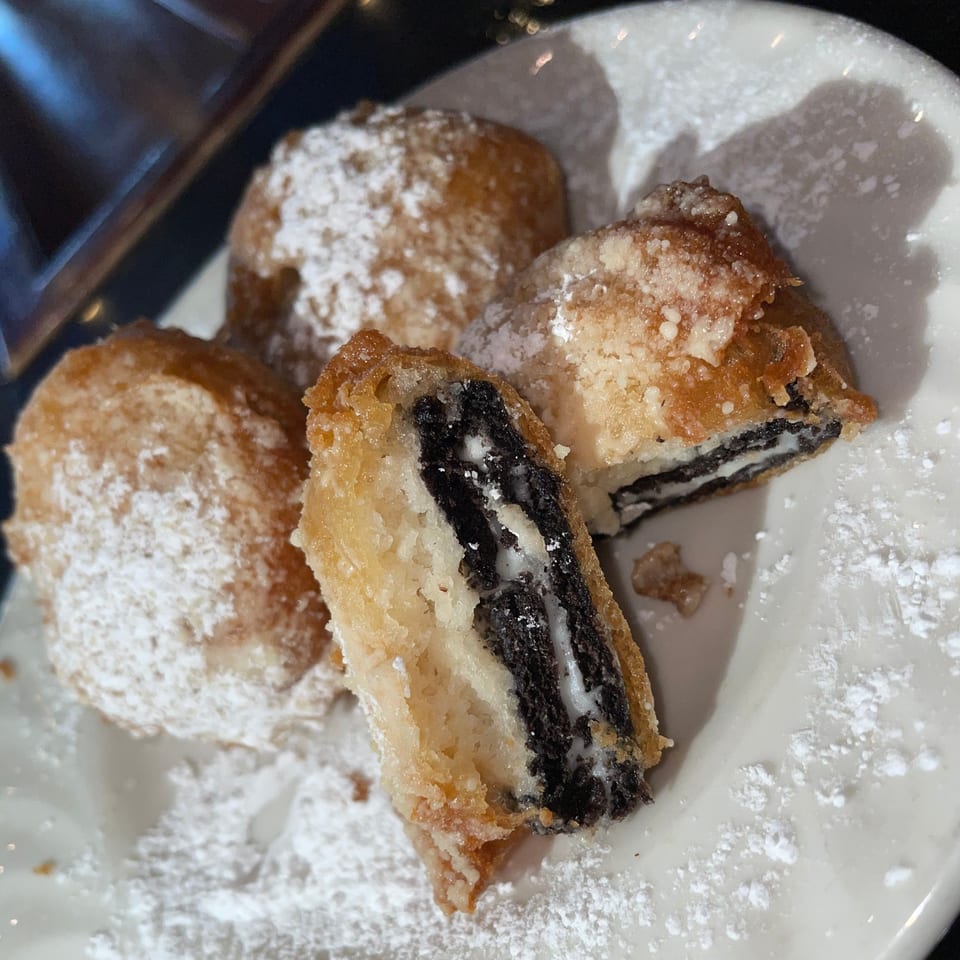 Four deep-fried Oreos dusted with powdered sugar on a white plate, one cut open revealing the cookie.