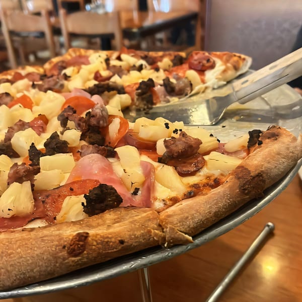 Close-up of a Homerun Pizza with pineapple, pepperoni, and sausage, with a server lifting a slice.