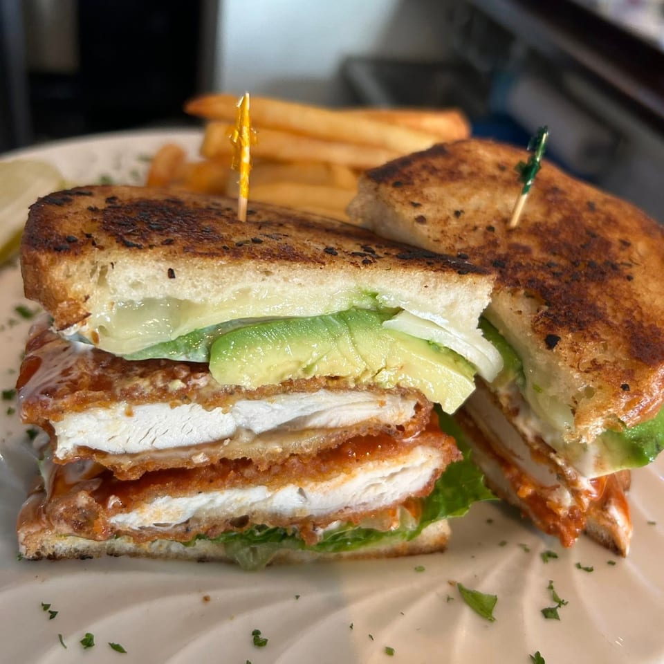 A crispy chicken sandwich with avocado, lettuce, and sauce, served with fries on a white plate.